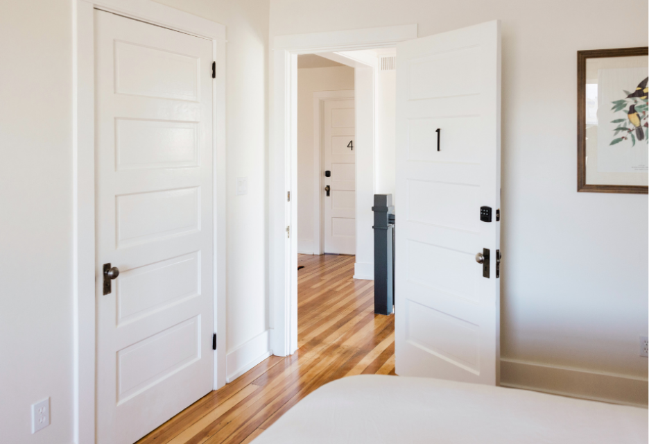 picture bedroom doorway taken form inside the bedroom over the corner of the bed.  the room has white walls and a brown polished hard wood floor. doorway leads into a hallway with the same color doors, walls and floors. 